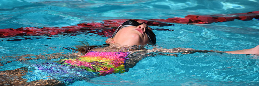 一个十几岁的女孩戴着护目镜和扎染泳衣在游泳池里仰泳