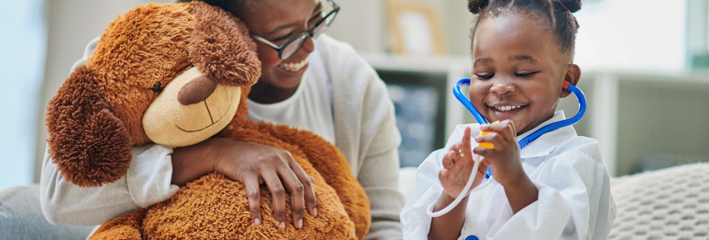 一个年轻的非洲裔美国女孩用玩具听诊器给一个填充动物做手术，她的妈妈坐在她旁边，笑着。