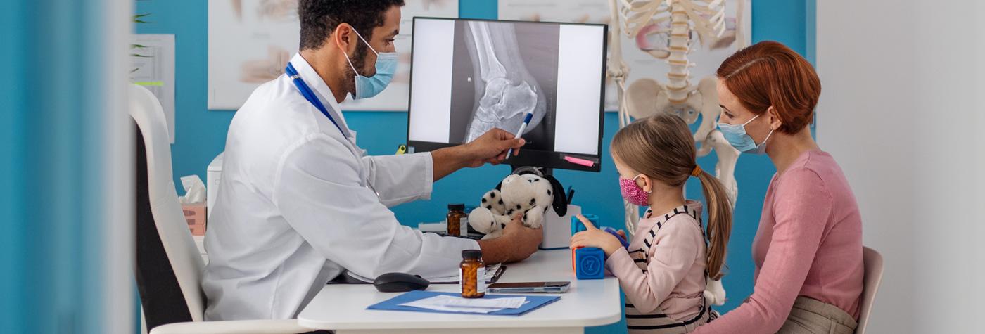 一个小女孩坐在医生办公桌前她妈妈的腿上，而医生手里拿着一个填充玩具，用一支笔指着电脑显示器上的x光图像。