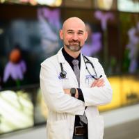 安德鲁·Kolodziej博士的照片微笑,摆姿势,双臂交叉在壁画前在医院。他是一个光头白人保管妥当的黑暗但胡须花白。他穿着一件白色外套黑色纽扣的衬衫,灰色的领带。