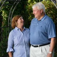 坦诚的照片E和她的丈夫对彼此微笑,站在一个金属网关在他们的花园。她的丈夫是一位年长的白人有白色的头发。他穿着一件蓝色的短袖马球,塞进他的卡其裤。